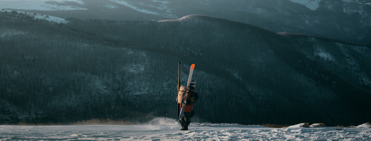 Person walking alone across a snowy landscape carrying skis on their back, with dark, forested mountains rising in the background.