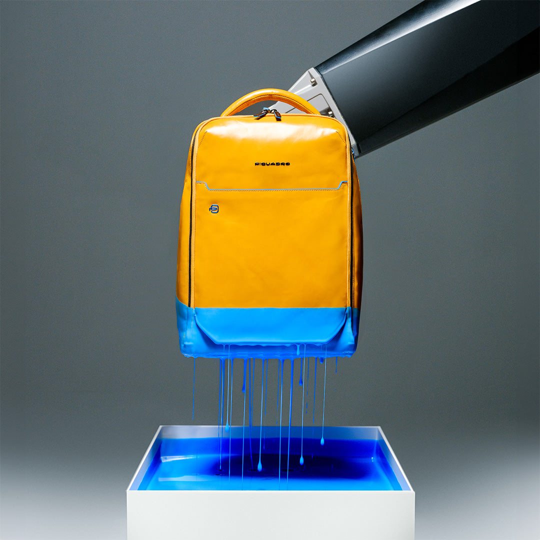 Yellow backpack being dipped into blue liquid, with blue paint dripping from the bottom into a square white container.